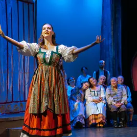 Performer in a long peasant dress singing with arms outstretched on a blue-lit stage, cast seated behind her