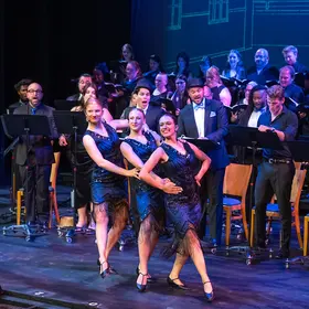 Dancers in dark dresses mid-step at the front of the stage with an ensemble and musicians behind