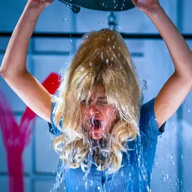 Performer in a big blonde wig with both arms raised overhead against a bright blue backdrop